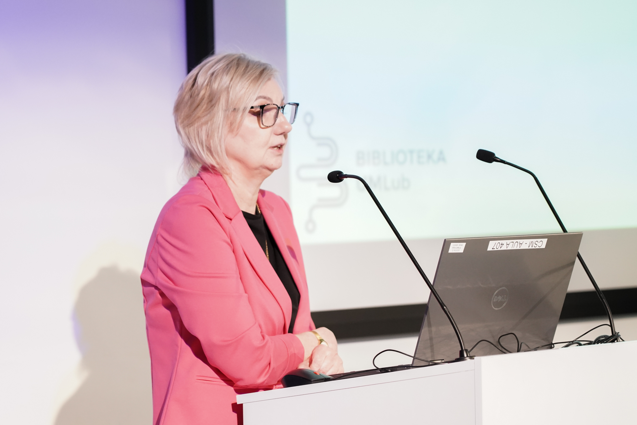 Elegancko ubrana kobieta za m&oacute;wnicą z dwoma mikrofonami, widziana z boku. Za nią wyświetla się napis biblioteka umlub